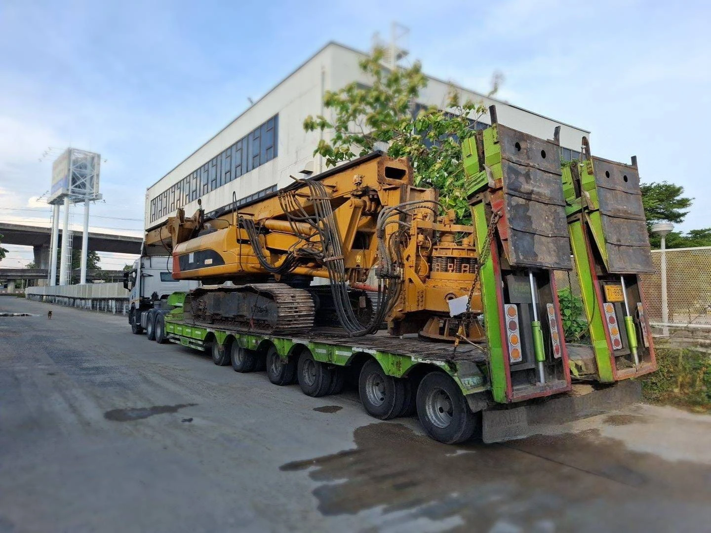 Heavy construction equipment on a long green trailer, carrying a large yellow excavator beside an industrial building on a road.